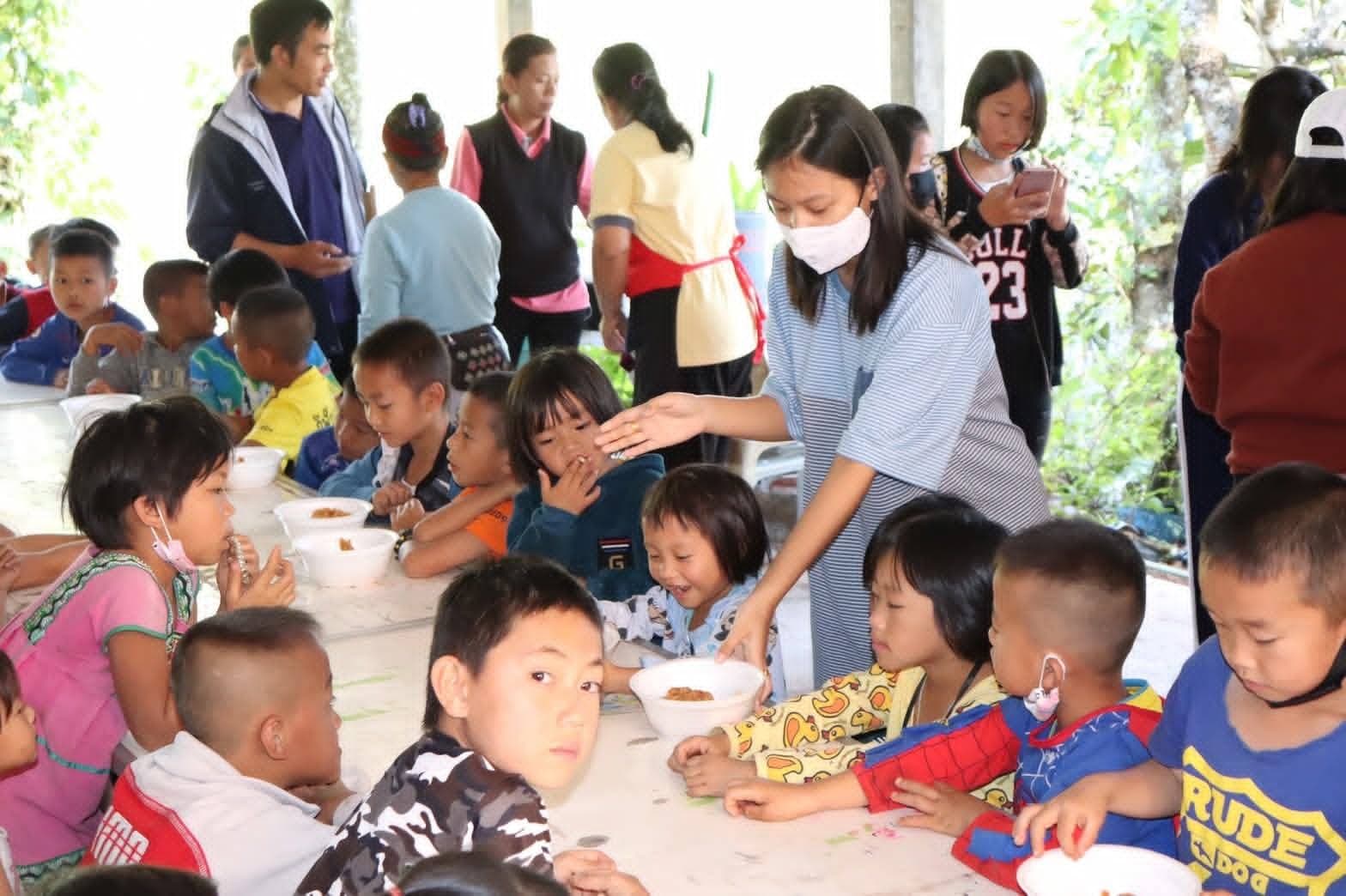 A volunteer placing a bowl of food for children seated at a long outdoor table